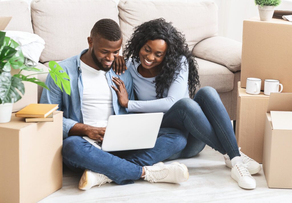 Just married African couple sitting on the floor of their new Las Vegas home, shopping online for furnishings using a laptop, embodying the simplicity and convenience of a streamlined rental search process. The empty space around them symbolizes the potential to personalize their living environment, facilitated by an efficient rental search in Las Vegas.