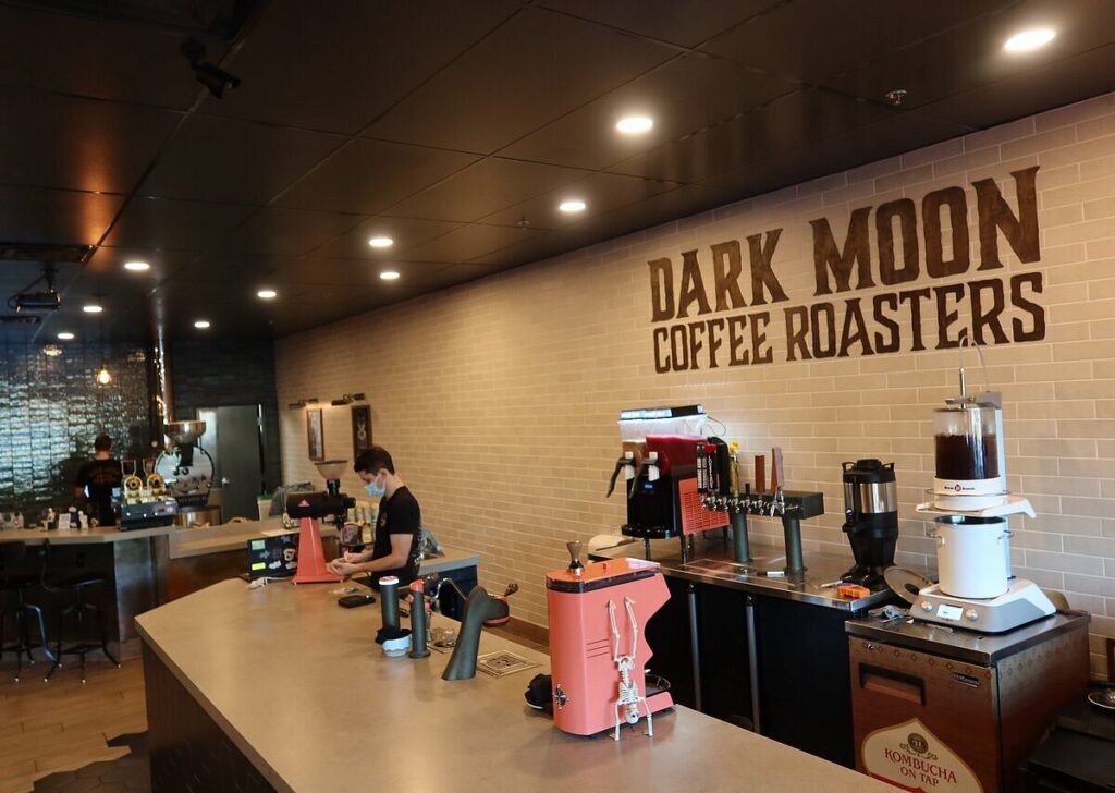 Interior of Dark Moon Coffee Roasters in Henderson showing a long service counter, high-end brewing equipment, and a large wall sign that reads "Dark Moon Coffee Roasters.