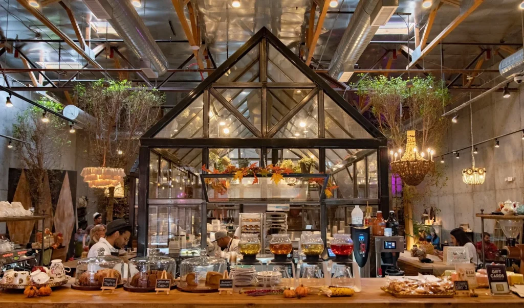 Interior view of Gäbi Coffee & Bakery in Las Vegas featuring a large glass greenhouse structure, industrial ceilings, and a rustic wooden counter with pastries. Caption: Architectural magic: The "house-within-a-house" glass structure that makes Gäbi a Chinatown icon.