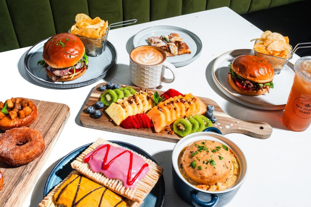 A high-angle flat lay of The Coffee Class menu items including house-made pop-tarts, gourmet burgers with chips, a fruit board, and various specialty lattes on a white table.