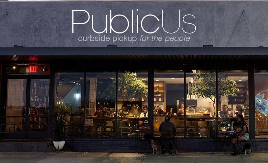 The exterior of PublicUs in Downtown Las Vegas at dusk, featuring a large sign that reads "PublicUs curbside pickup for the people" above large glass windows showing a warm interior with trees.