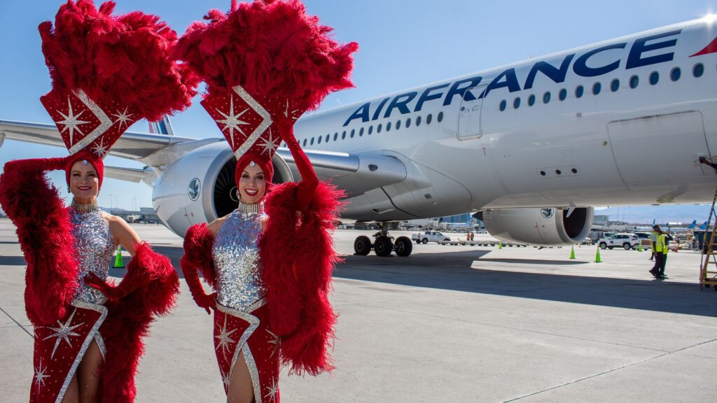 Two Las Vegas showgirls in glittering silver and red feather costumes posing in front of an inaugural Air France Airbus A350 on the tarmac at Harry Reid International Airport.