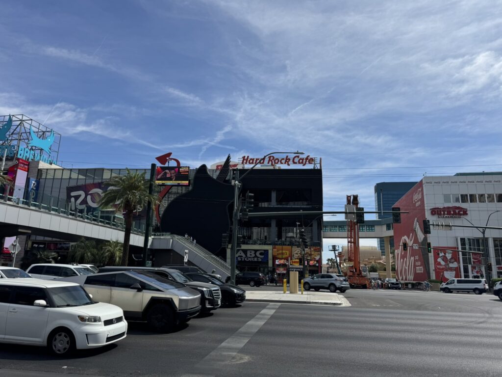 A wide daytime shot of the Las Vegas Strip showing the Hard Rock Cafe facade where the traditional neon guitar has been replaced by a large, matte-black LED screen shaped like a guitar body.