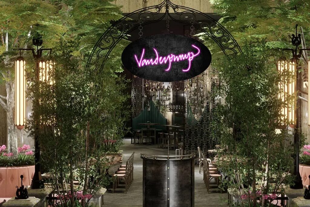 A lush, green entryway at The Vanderpump Hotel in Las Vegas featuring a black wrought-iron archway and a glowing pink neon "Vanderpump" sign.