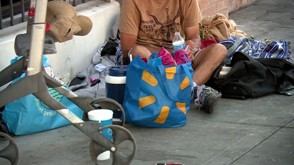 An unhoused individual sitting on a downtown Las Vegas sidewalk next to a blue Walmart bag, blankets, and a walker, representing the local street community.