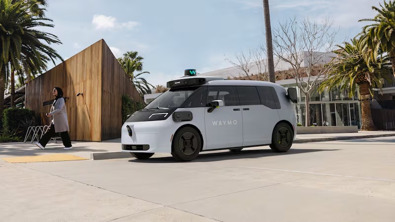 A white, fully autonomous Waymo robotaxi vehicle parked on a paved street in front of modern buildings and palm trees.
